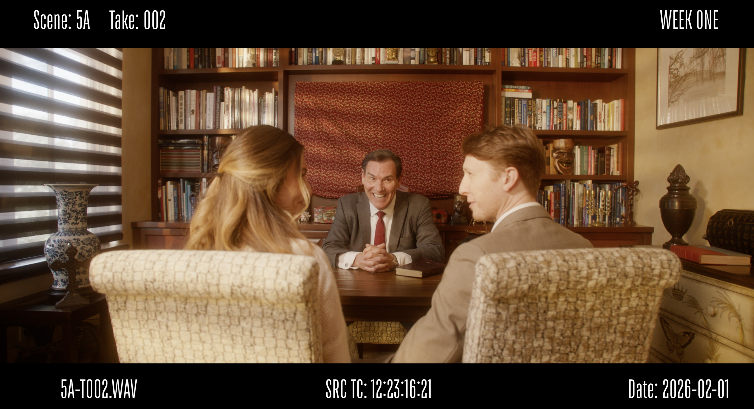 A scene of four people sitting around a table in a cozy library or study, with shelves of books behind them. One man in a suit is smiling at two others, a woman and a man, who are sitting opposite him.