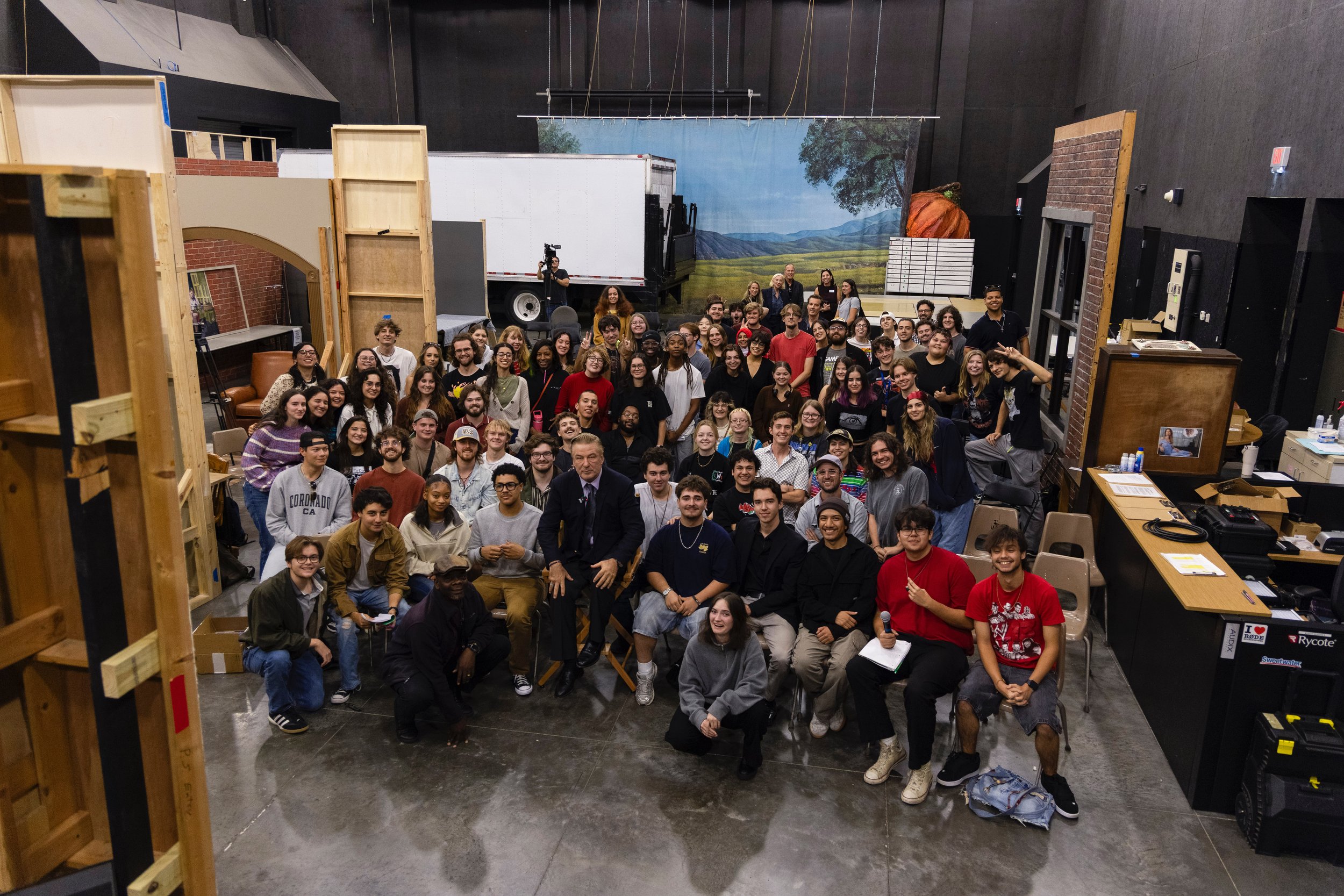 Group photo of diverse people in a large indoor space, some smiling and looking at the camera, with a painted mural of a landscape in the background and a person holding a camera.