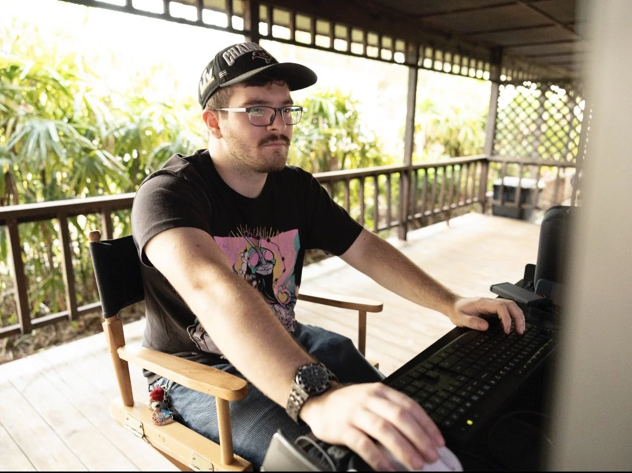 Man with glasses, baseball cap, and black T-shirt with colorful graphic, working on a computer at a wooden desk on a porch with greenery in the background.