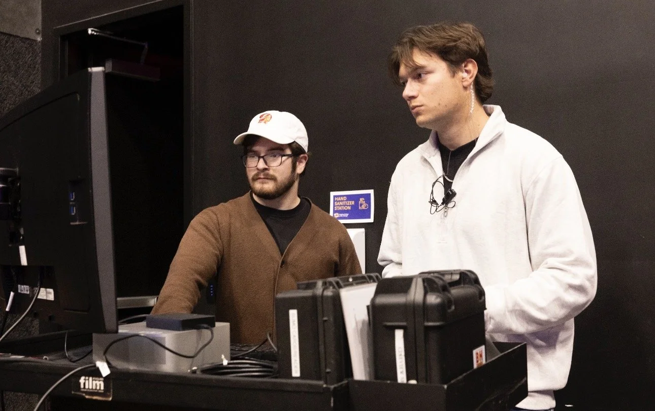 Two men standing in front of a computer and filming equipment, one wearing a white jacket and the other wearing glasses, a brown sweater, and a baseball cap, in an indoor setting with a black wall background.