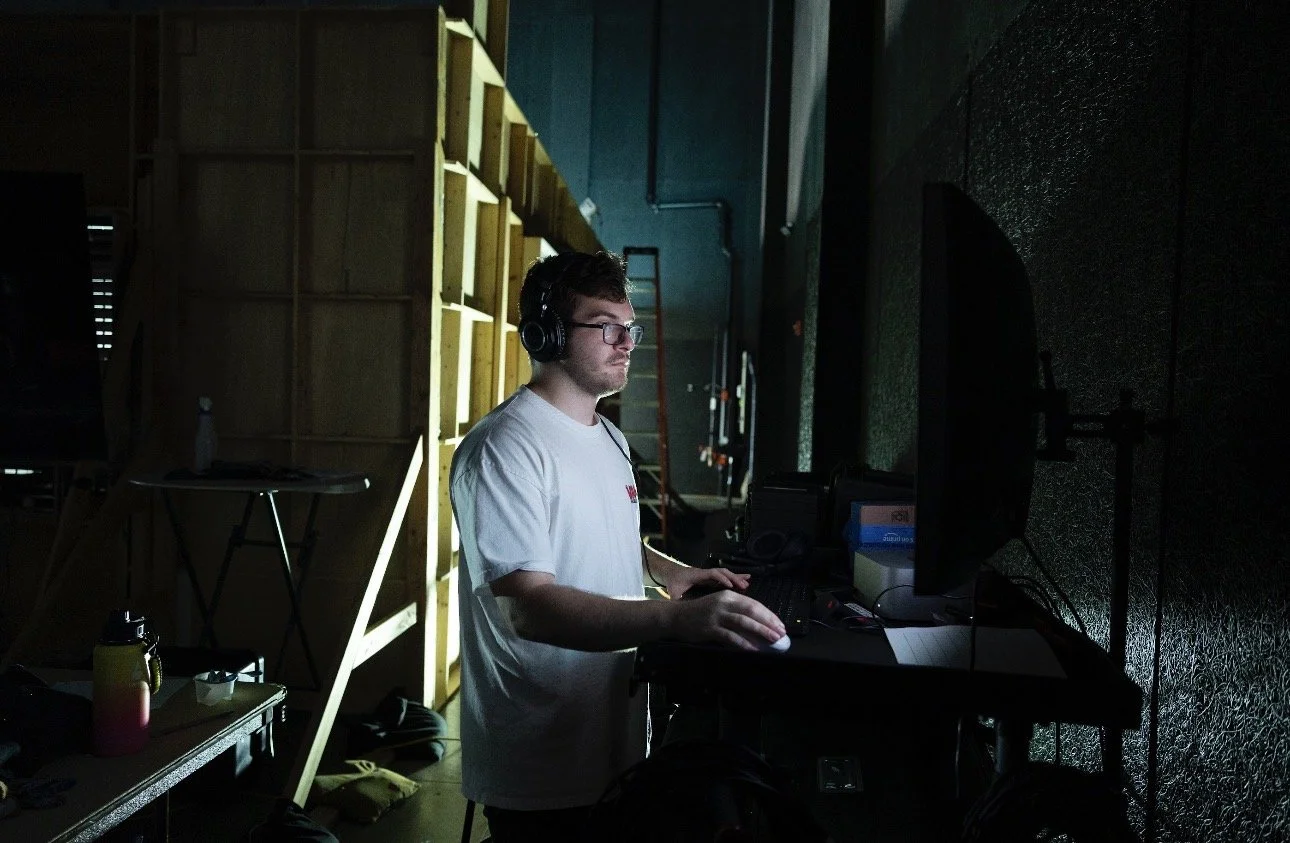 A man wearing glasses and headphones, working on a computer in a dimly lit room with soundproofing panels on the wall.