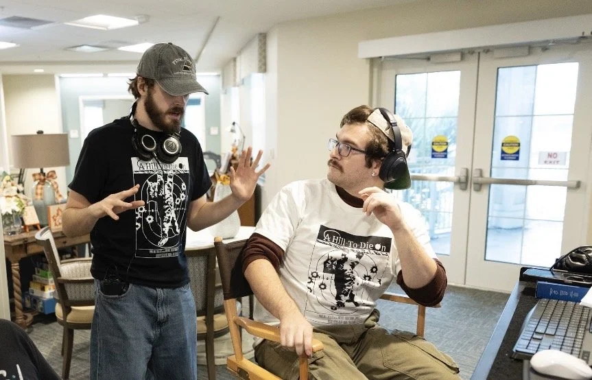 Two young men wearing headphones are engaged in conversation in a room with glass doors, a table, and chairs.
