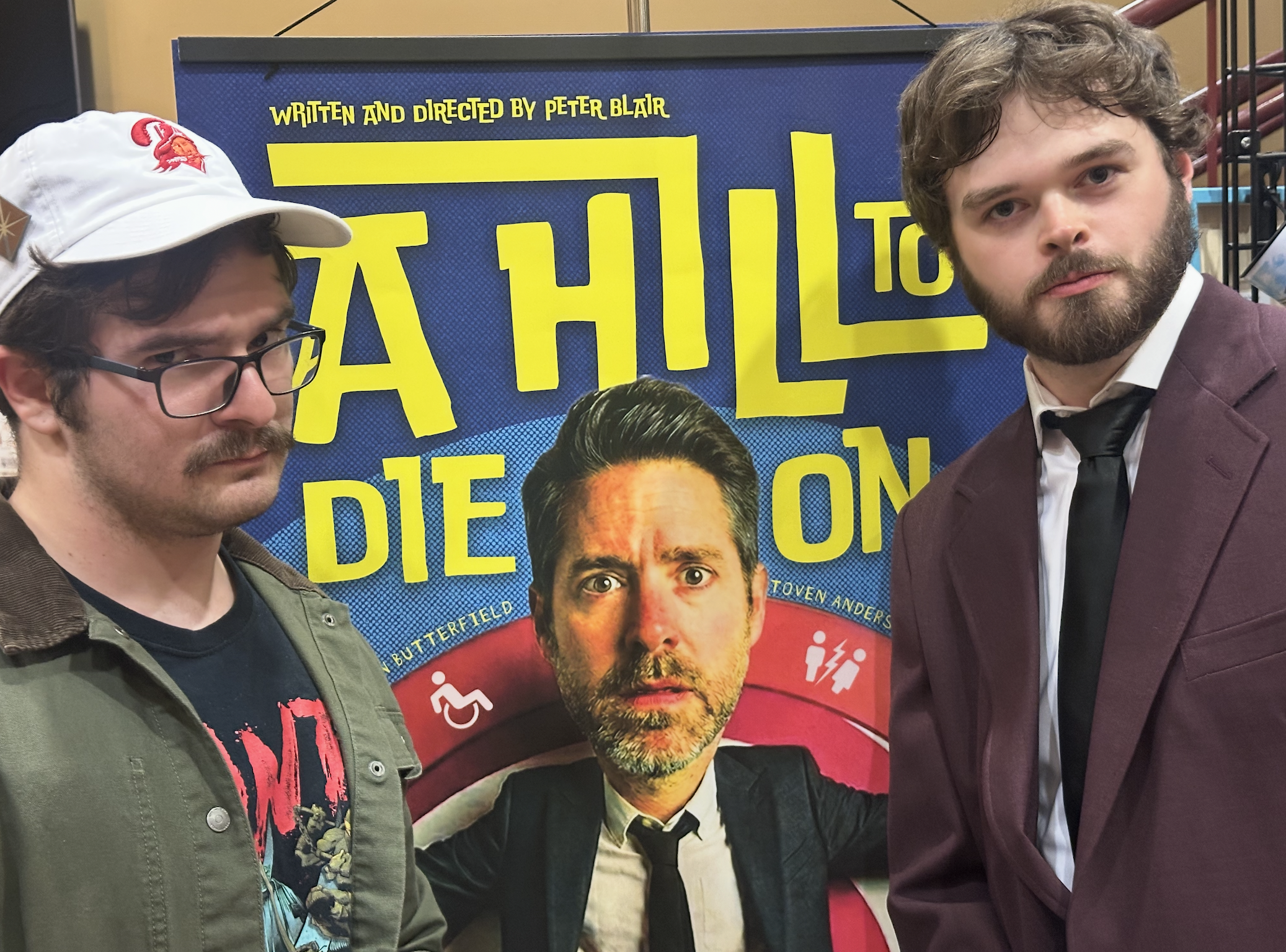 Two men stand in front of a colorful poster featuring a portrait of a man with a beard and mustache, and text indicating it is written and directed by Peter Blair. The man on the left has dark hair, glasses, a mustache, and wears a green jacket, whil
