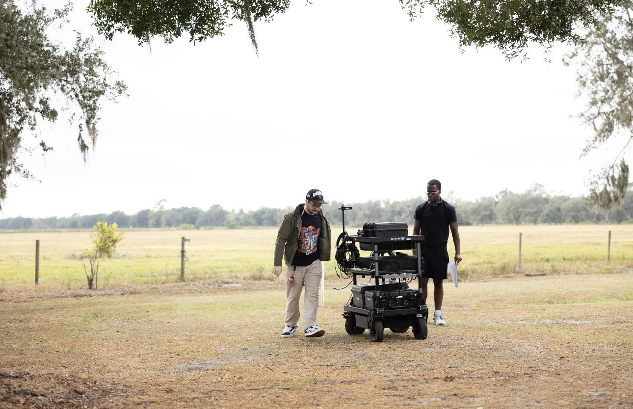 Two people on a film set outdoors with equipment, surrounded by open fields and trees overhead.
