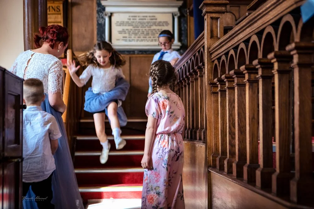 Children dressed in formal attire playing and interacting on a staircase inside a building, possibly a church, with wooden railings and a framed document or sign in the background.
