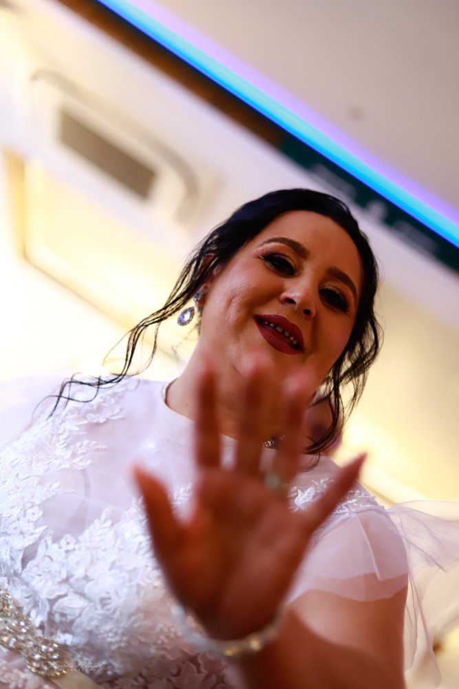 A woman with dark hair, makeup, and earrings is raising her hand toward the camera, smiling, in a well-lit indoor setting, wearing a white lace dress.