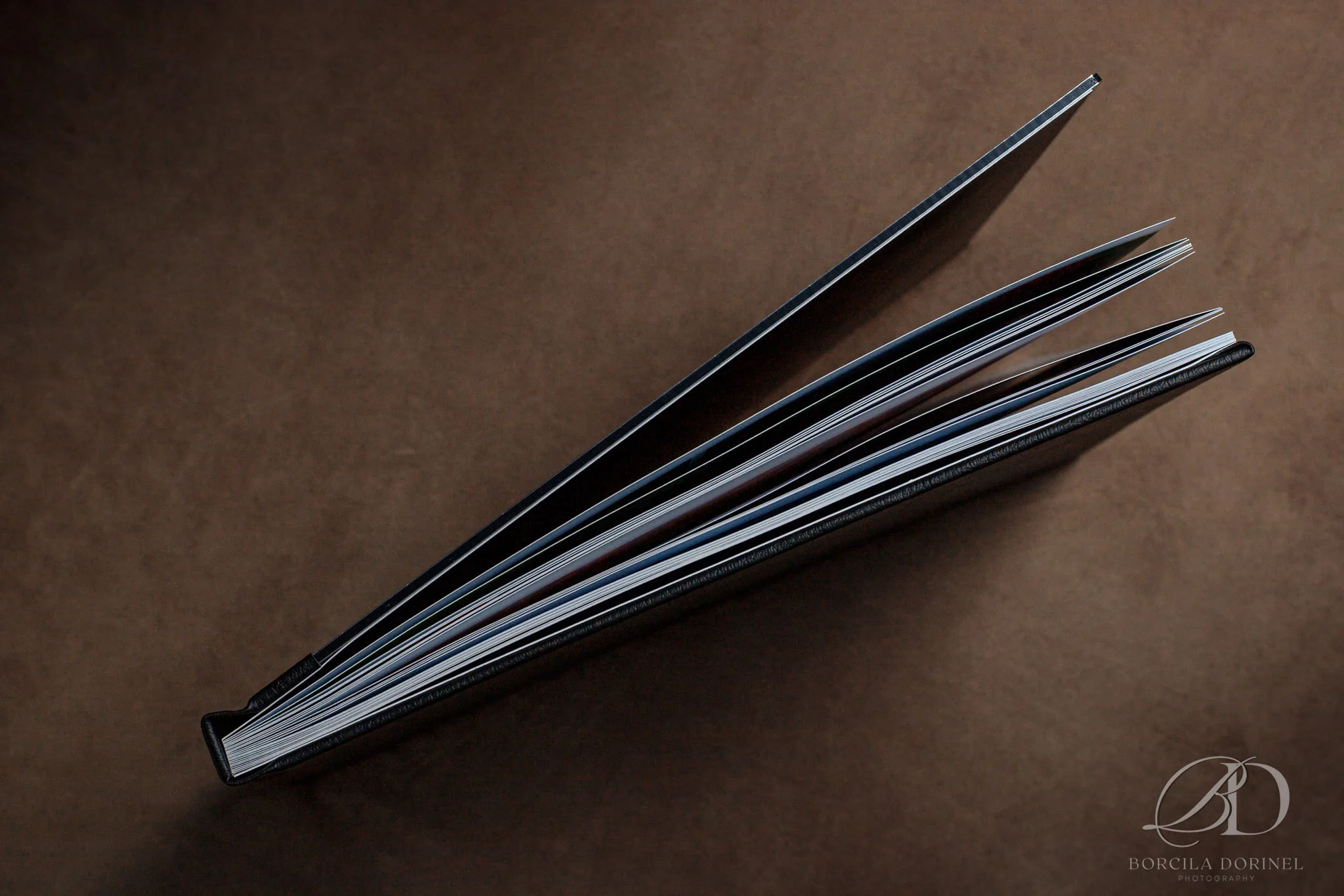 Top view of an open black hardcover book with pages fanned out on a brown textured background.