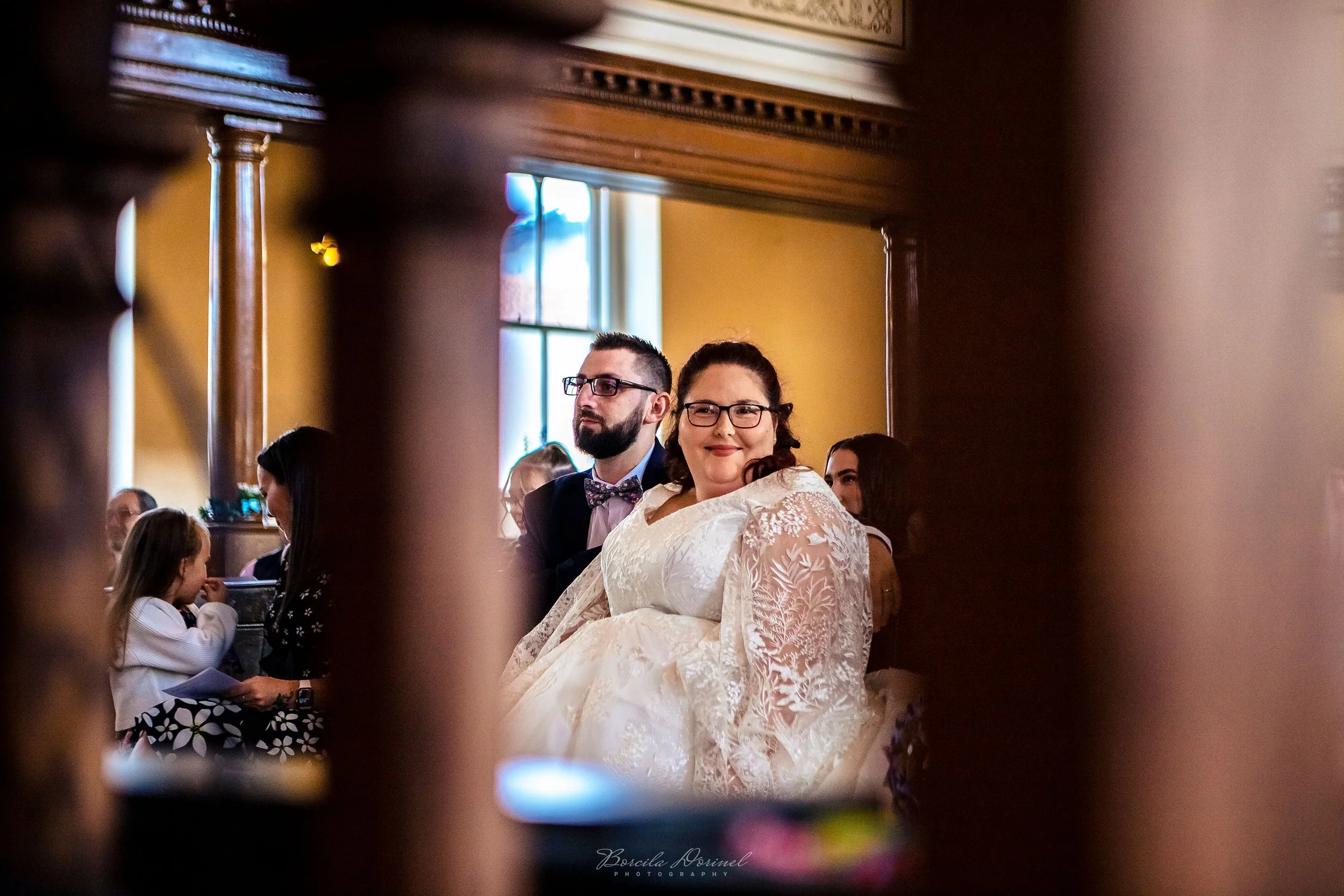A woman in a white lace dress and glasses smiling at a formal event, seated next to a bearded man in glasses, suit, and bow tie, in a warmly lit room seen through a wooden railing.