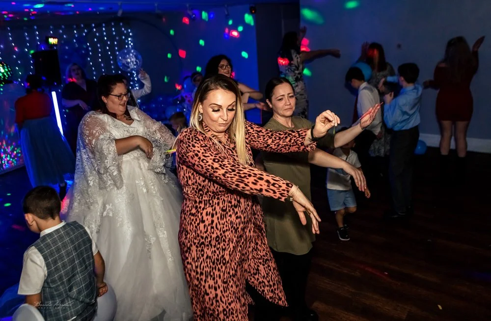 People dancing at a party with colorful lights and decorations, including a woman in a floral dress and others in formal and casual attire.