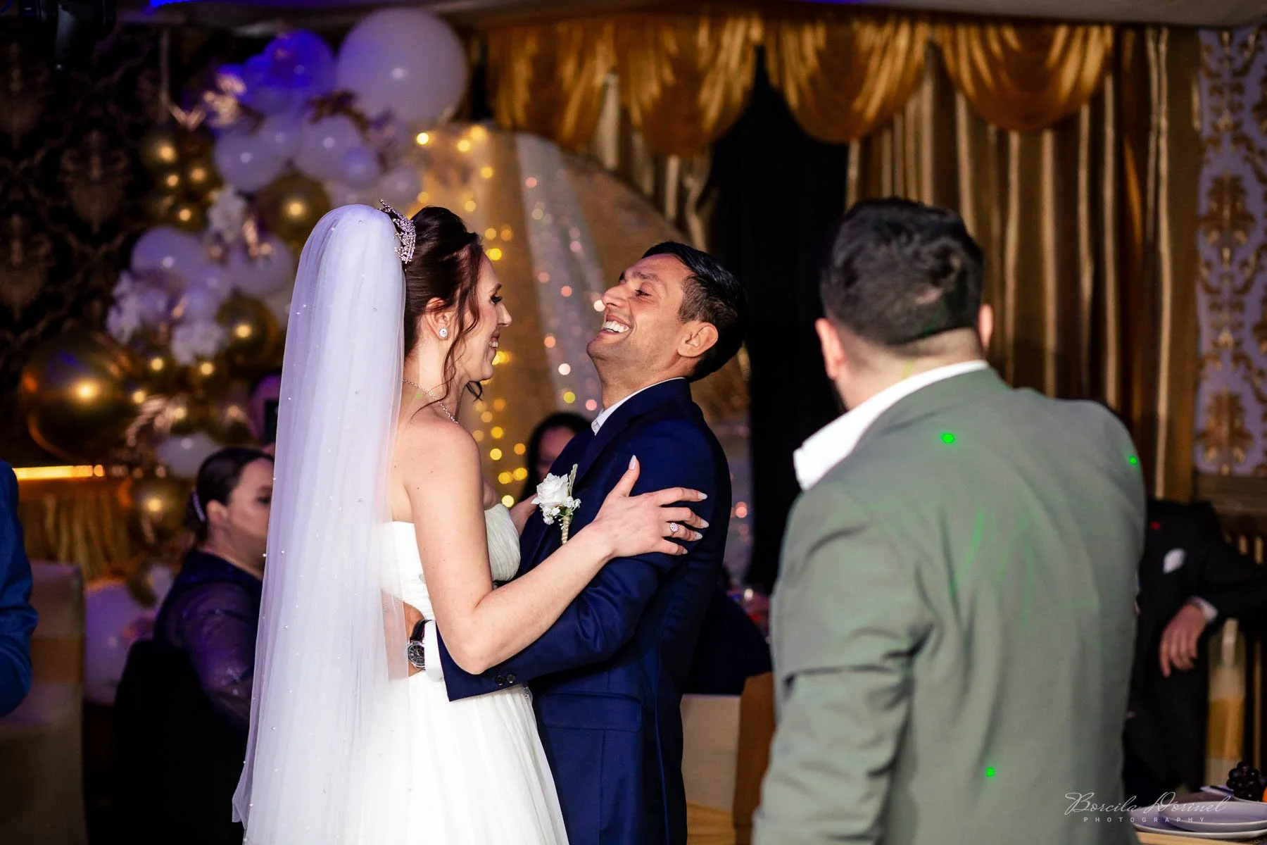 A bride and groom holding each other and smiling during their wedding celebration, with a background of balloons, golden curtains, and guests.