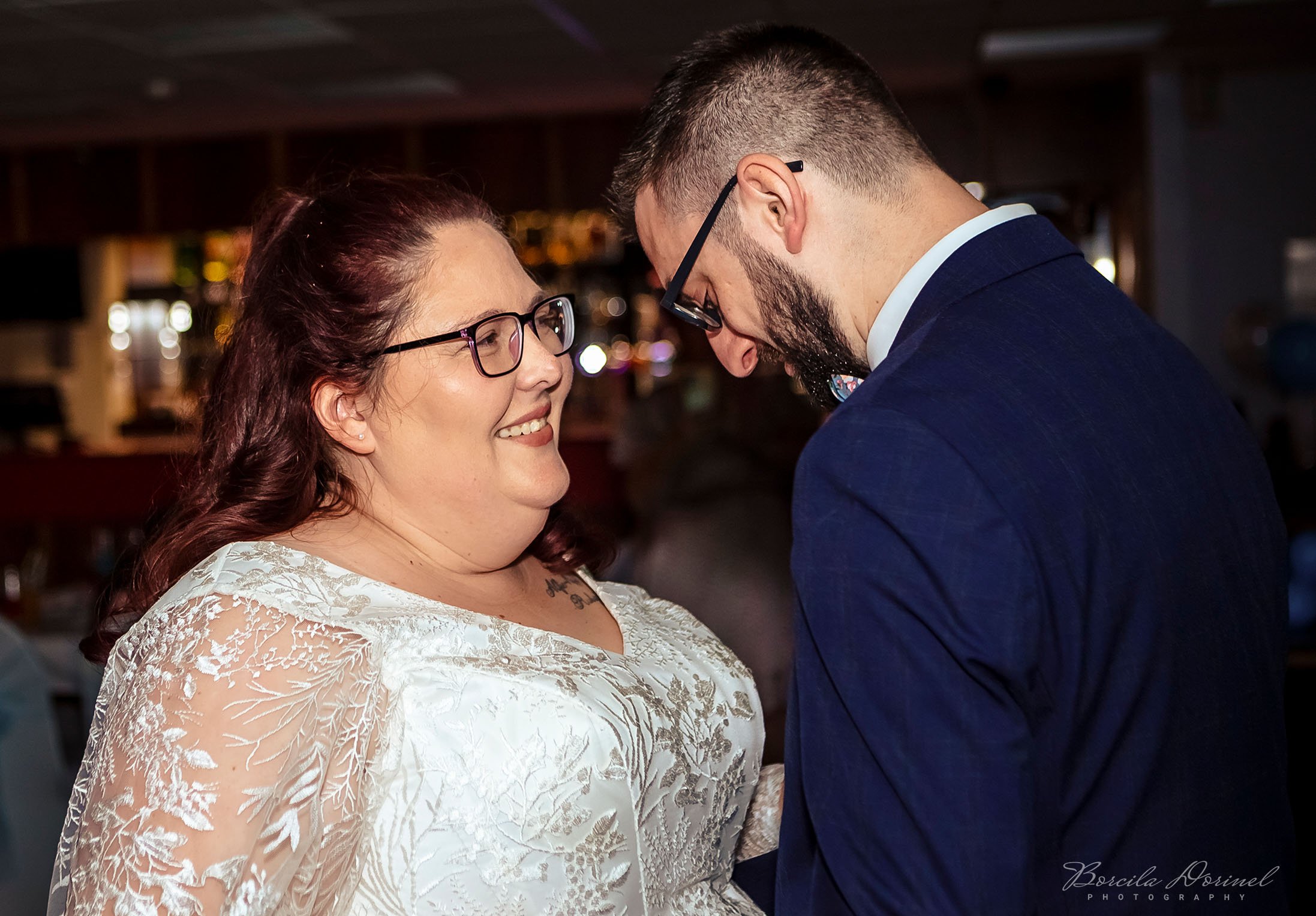 A couple dancing closely, smiling, with the woman wearing glasses and a white lace dress, and the man wearing glasses and a dark blue suit.