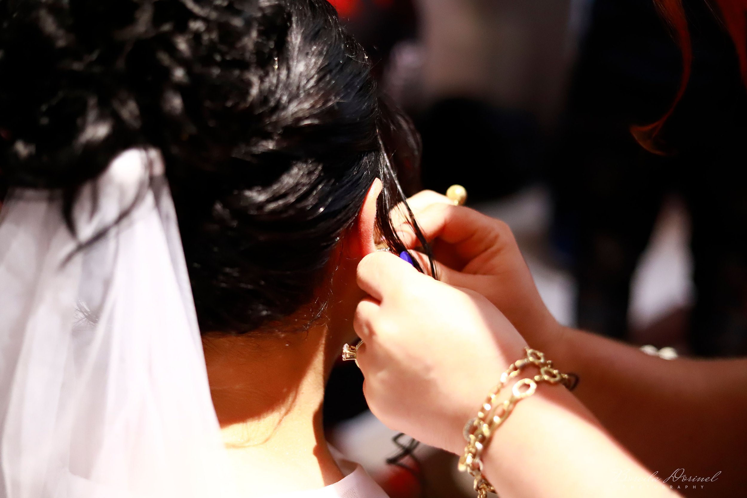 A close-up of a woman’s ear being adorned with an earring during a special event, with a hand holding the earring and wearing a gold bracelet.