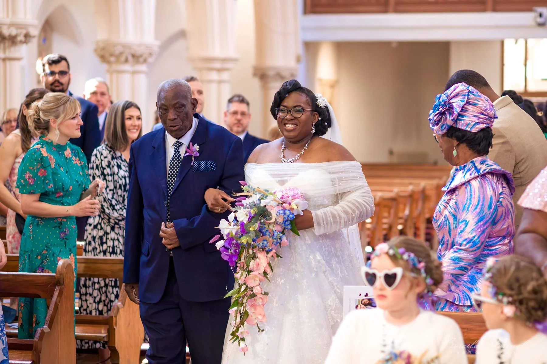 A bride with glasses, smiling and holding a floral bouquet, walking down the aisle with a man in a navy suit, surrounded by seated and standing wedding guests in a church.
