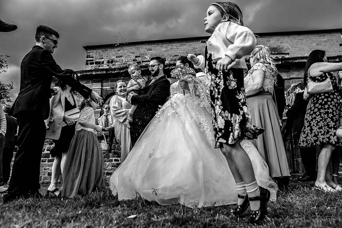 Black and white photo of a wedding celebration with guests dancing outdoors near a brick building, including children and adults dressed in formal attire.