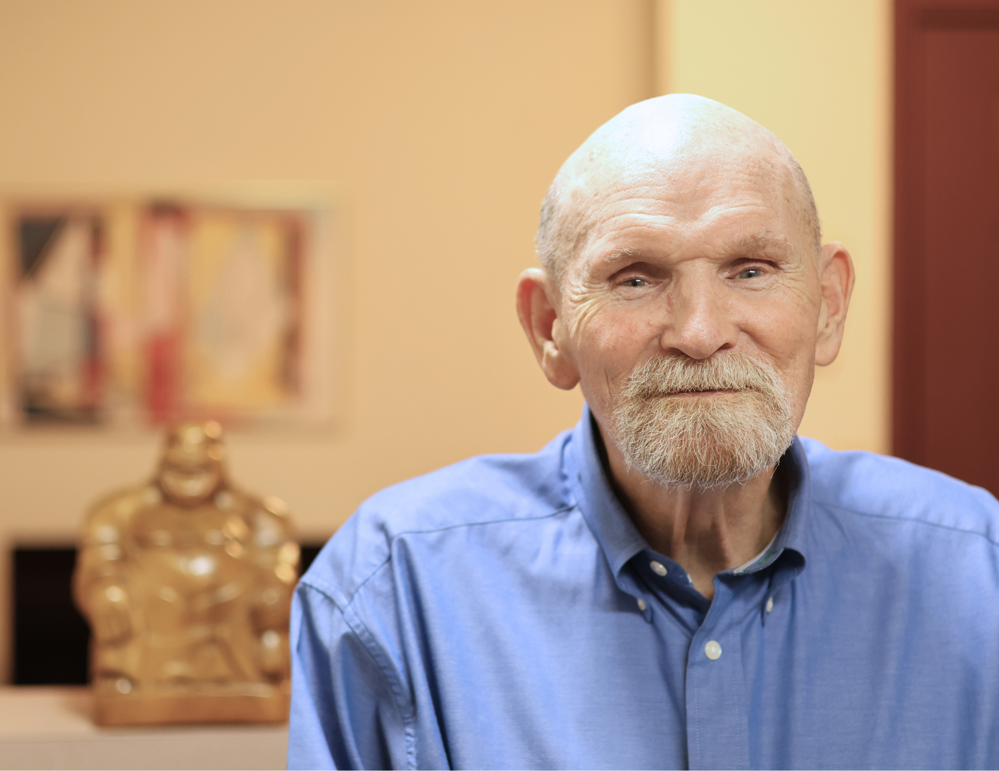 A smiling elderly man with a beard and mustache wearing a blue button-up shirt, sitting in an indoor setting with artwork and a decorative sculpture in the background.