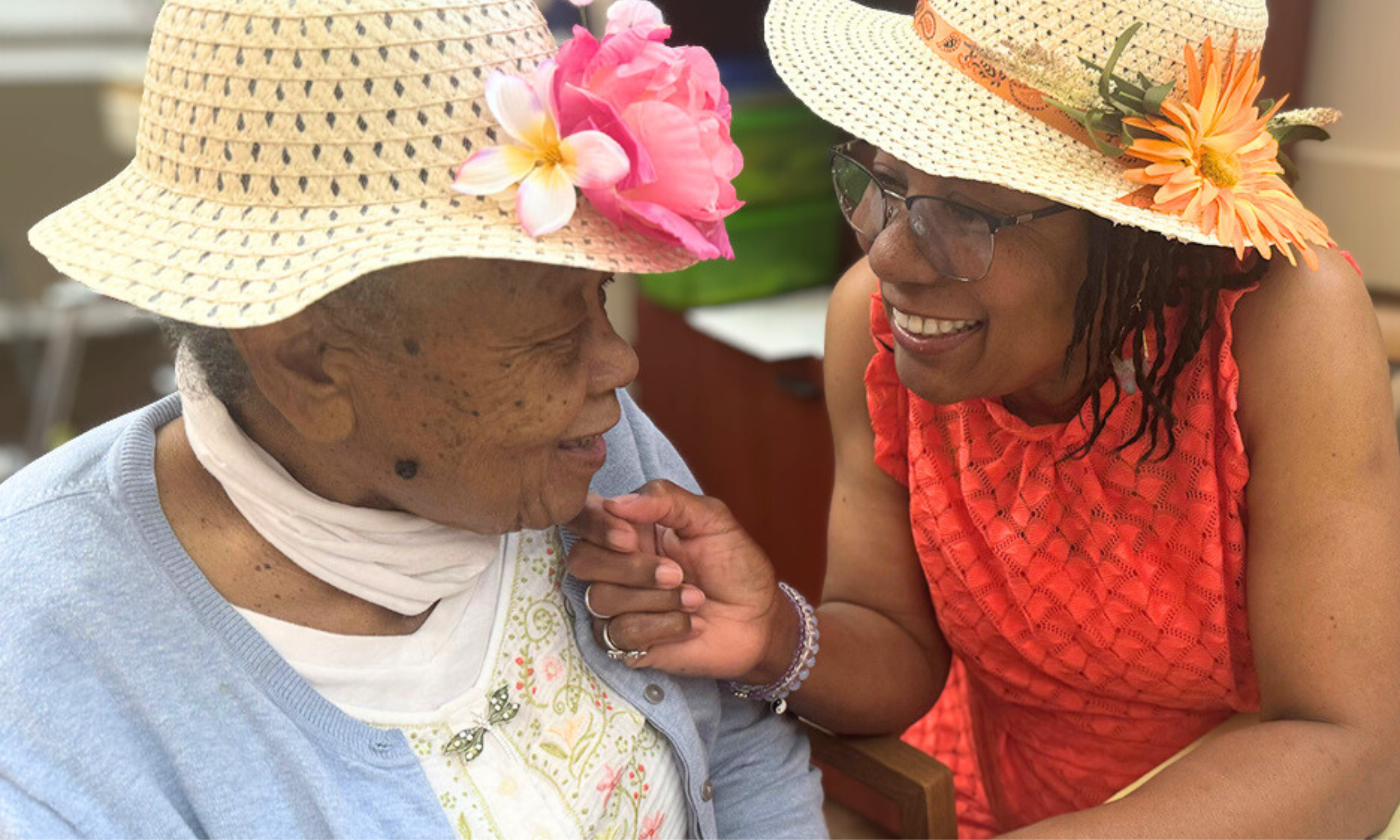 Two women wearing floral straw hats and smiling at each other, sharing a happy moment.