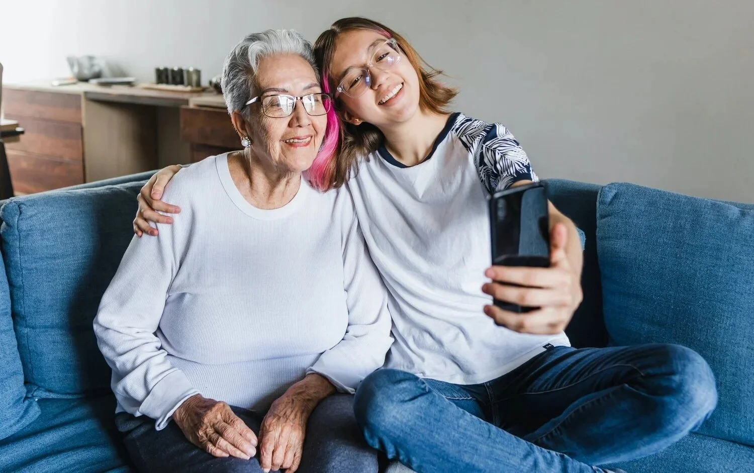 A young woman and an elderly woman taking a selfie together on a blue couch, smiling and hugging.