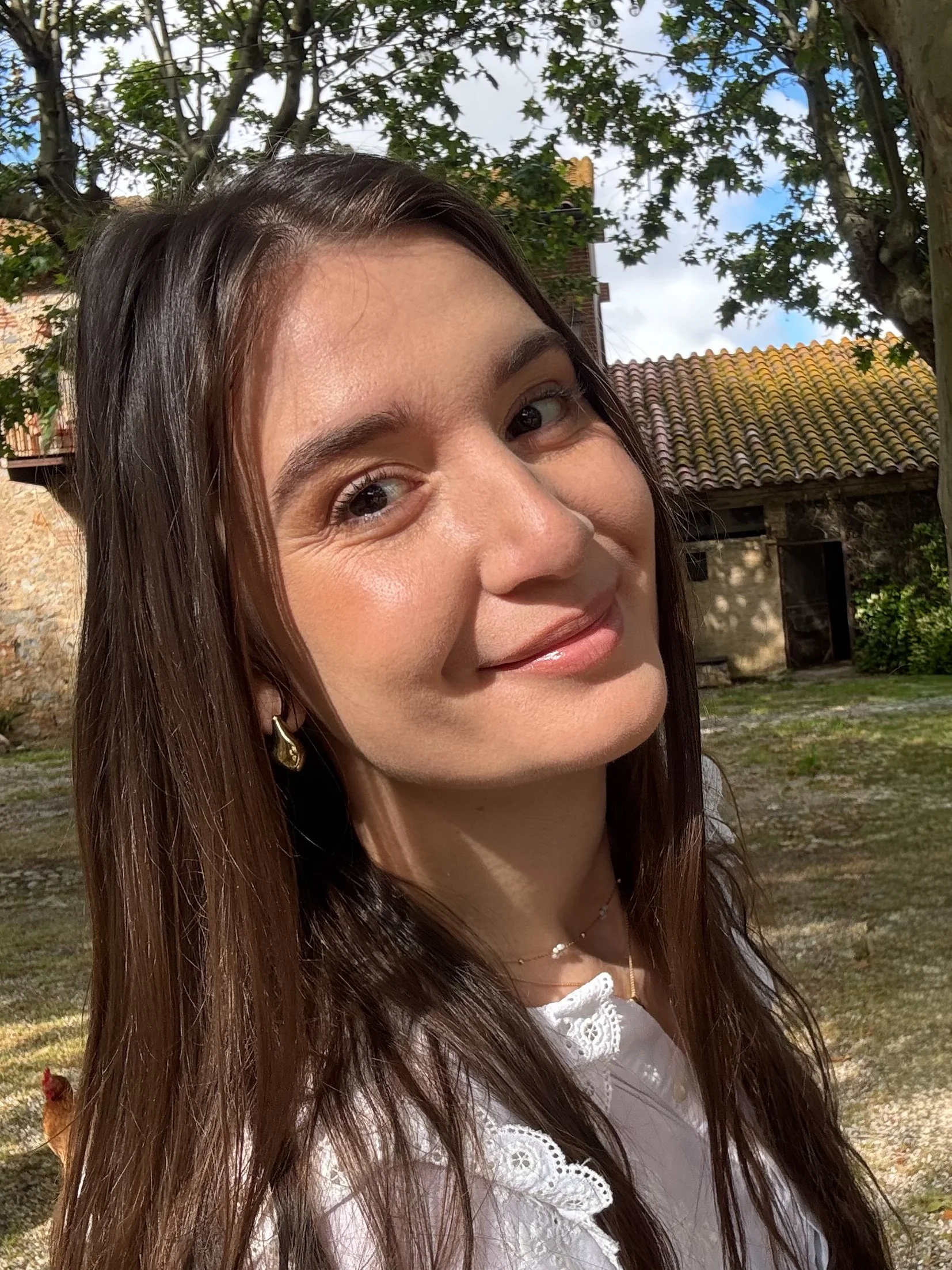 Jeune femme avec longs cheveux bruns, souriant, portant une boucle d'oreille en forme de goutte et une chaîne délicate, en extérieur avec des arbres et un bâtiment en arrière-plan, ciel partiellement nuageux. Maquillage très naturel et lumineux