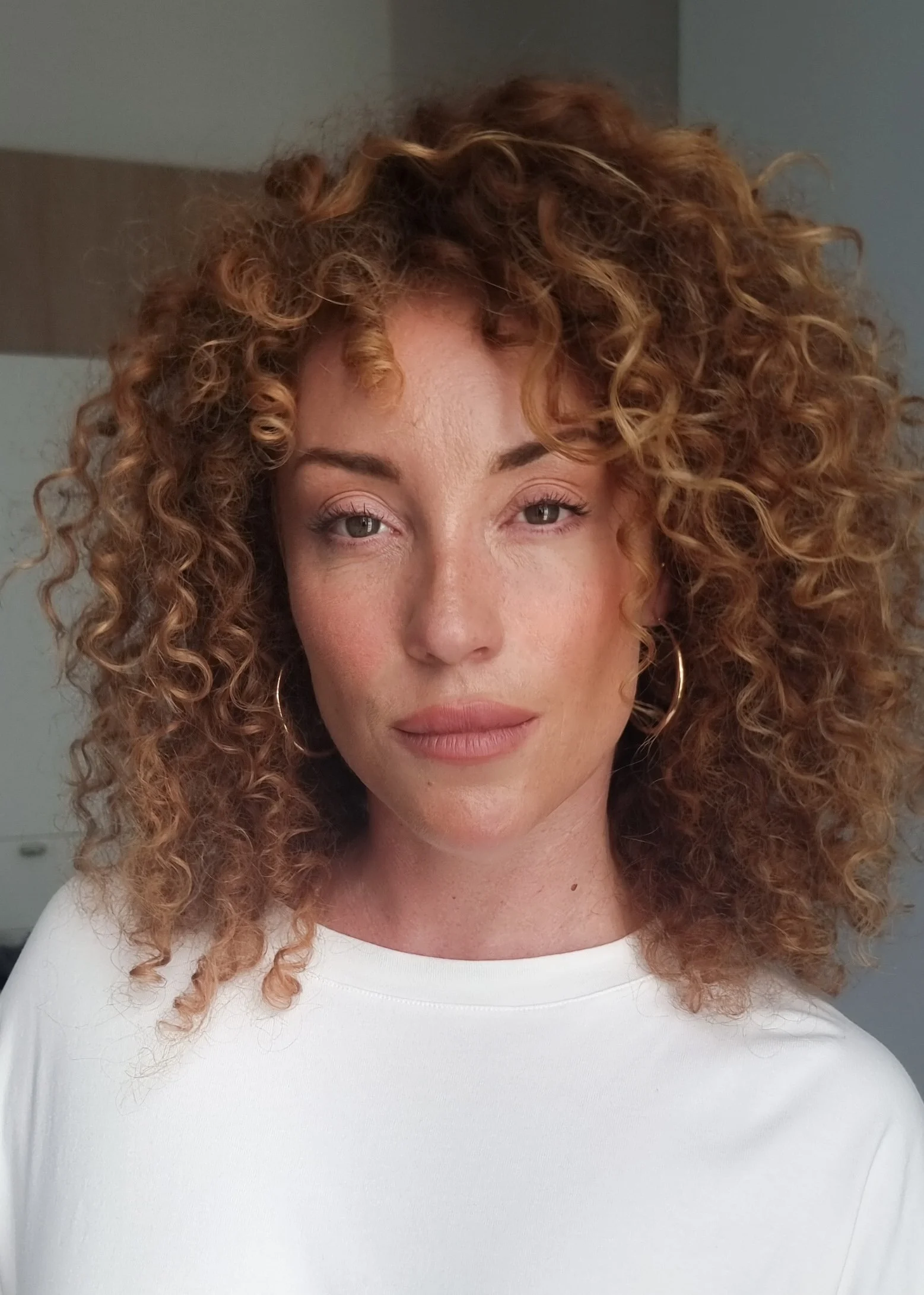 Portrait d'une femme avec des cheveux roux bouclés portant un tshirt blanc, regardant vers l'avant dans une pièce intérieure.