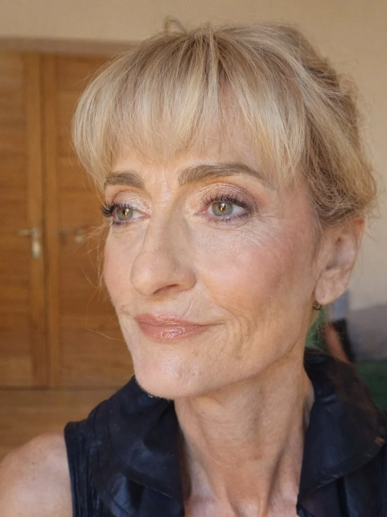 Une femme âgée souriante avec des cheveux blonds avec un maquillage sophistiqué et élégant , portant un haut noir, dans un intérieur avec un fond en bois.