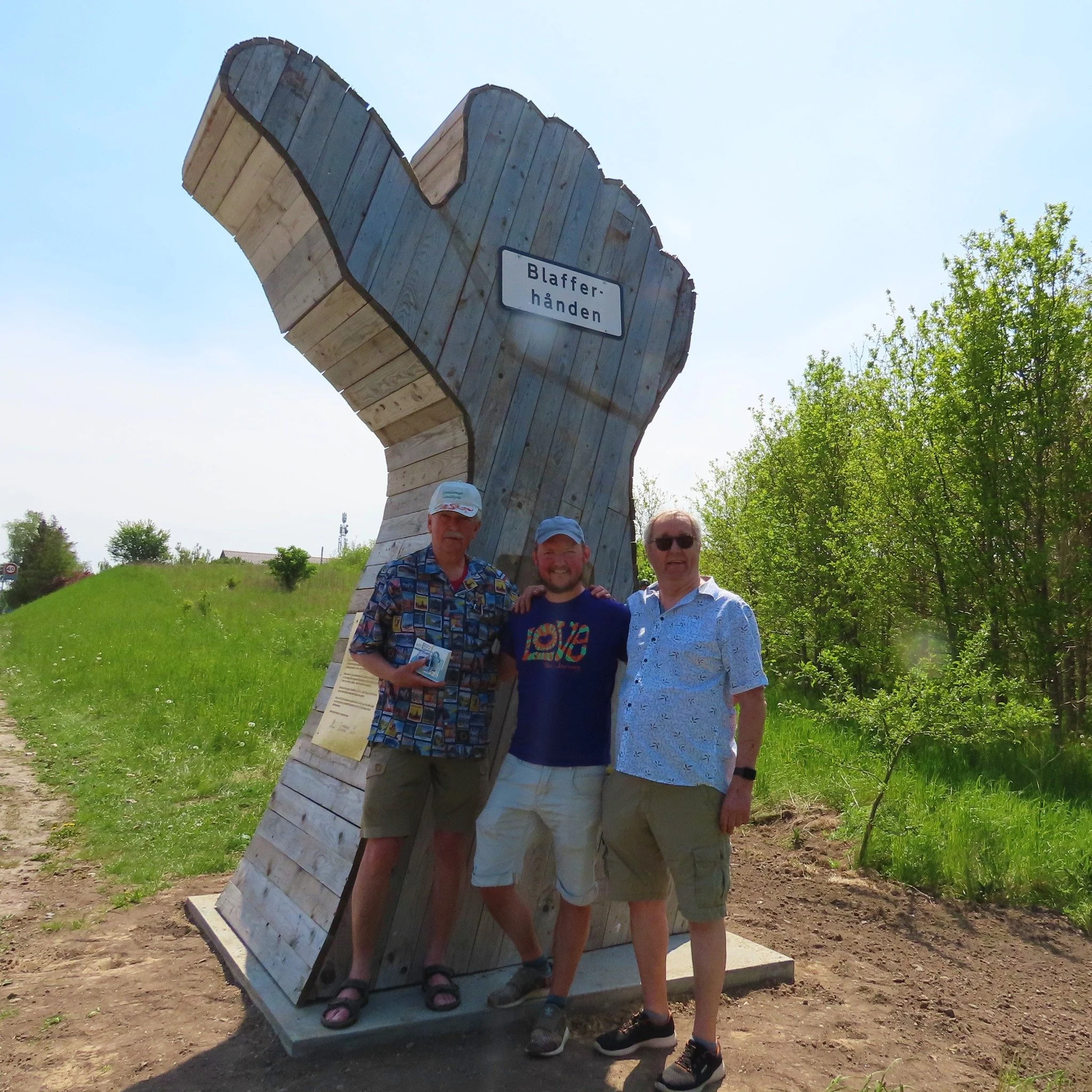 Tre personer står foran en stor, kunstnerisk skulptur af en hændelig hånd med en skilt, der siger 'Blaffer-hånden', på en græsmark med træer i baggrunden.