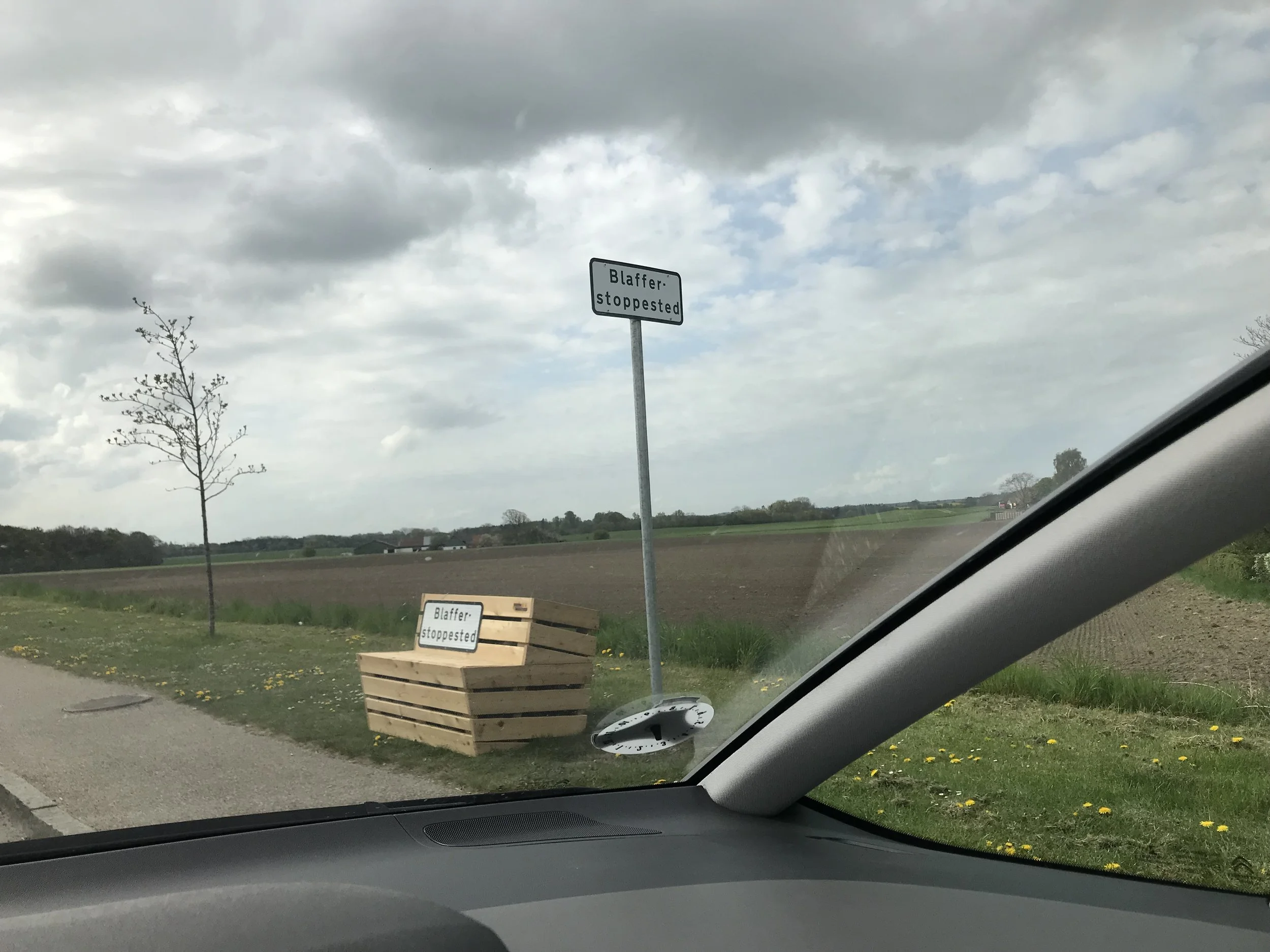 Et stopskilt med teksten 'Blaffer stoppested' samt en række træbænke med et lignende skilt på, ved siden af en landevej med landbrugsjord i baggrunden.