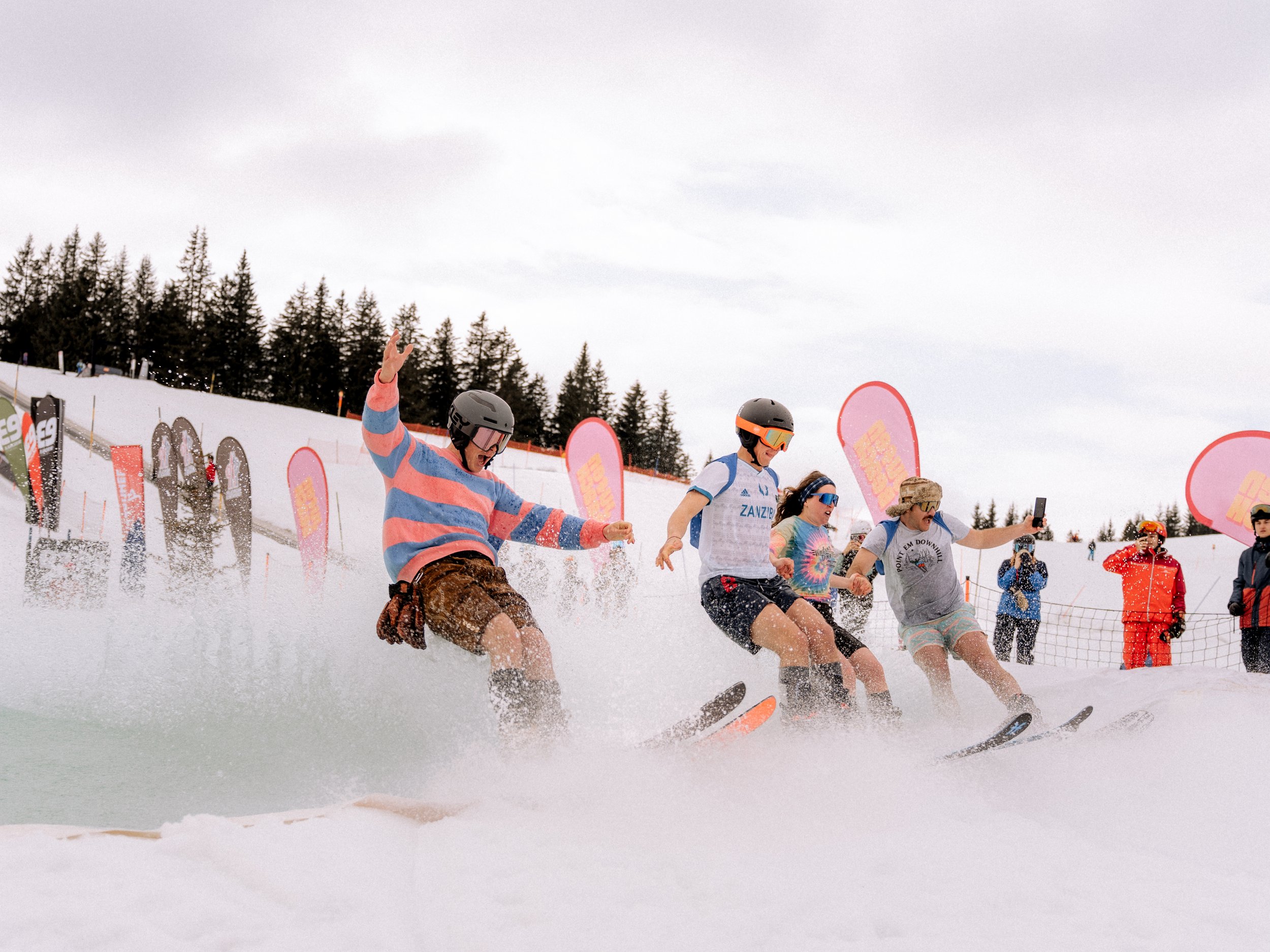 Mehrere Personen mit Skiern fahren durch Wasser auf einer schneebedeckten Piste. Einige tragen Helme und bunte Kleidung. Im Hintergrund sind Bäume und bunte Fahnen zu sehen.