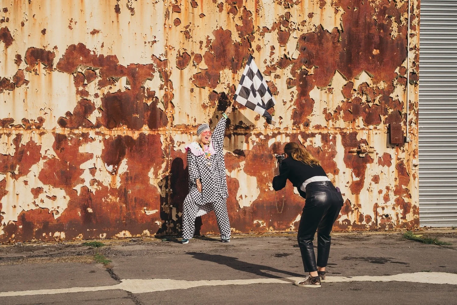 A woman influencer holding a checkered flag, being photographed by another woman against a rusted metal wall at a She's Electric on track event with Alfa Romeo