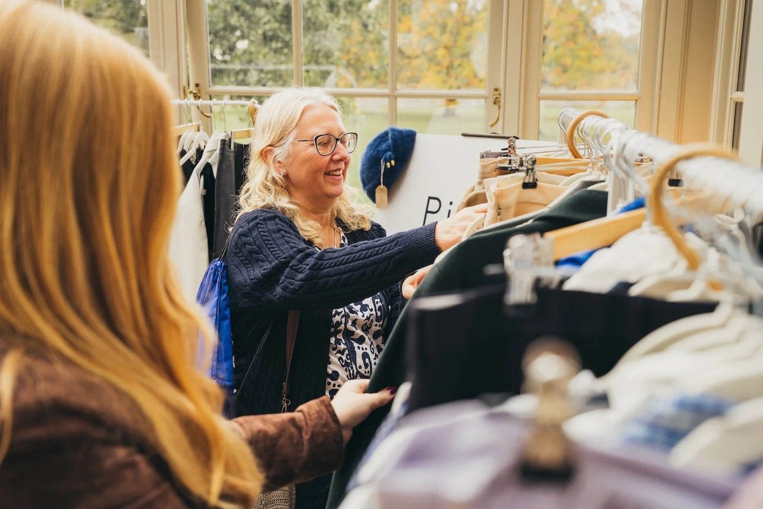 Two women shopping for clothes at an indoor thrift store or secondhand shop, browsing through a rack of clothing.