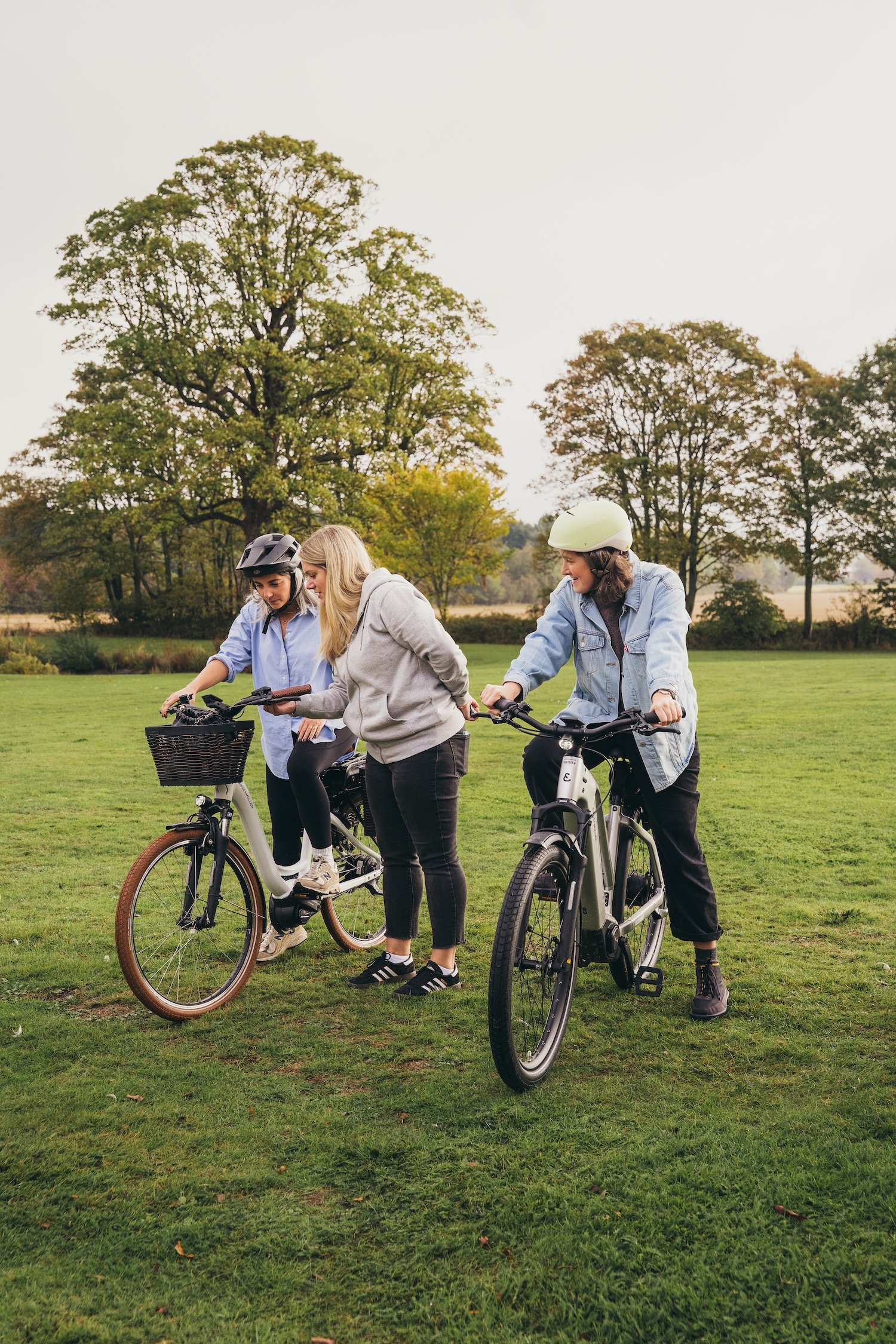 Three women with helmets and casual clothing standing and riding electric bikes in a park with green grass and trees.