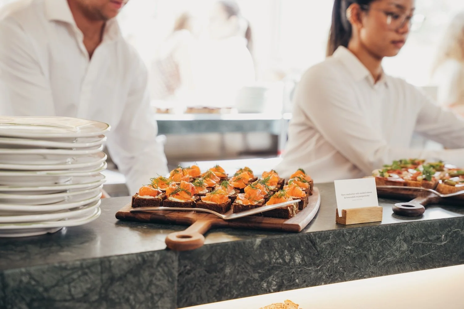 Appetizer tray with smoked salmon on dark bread, garnished with herbs, at a buffet table. Stacks of white plates are on the left, and a woman is serving herself in the background.