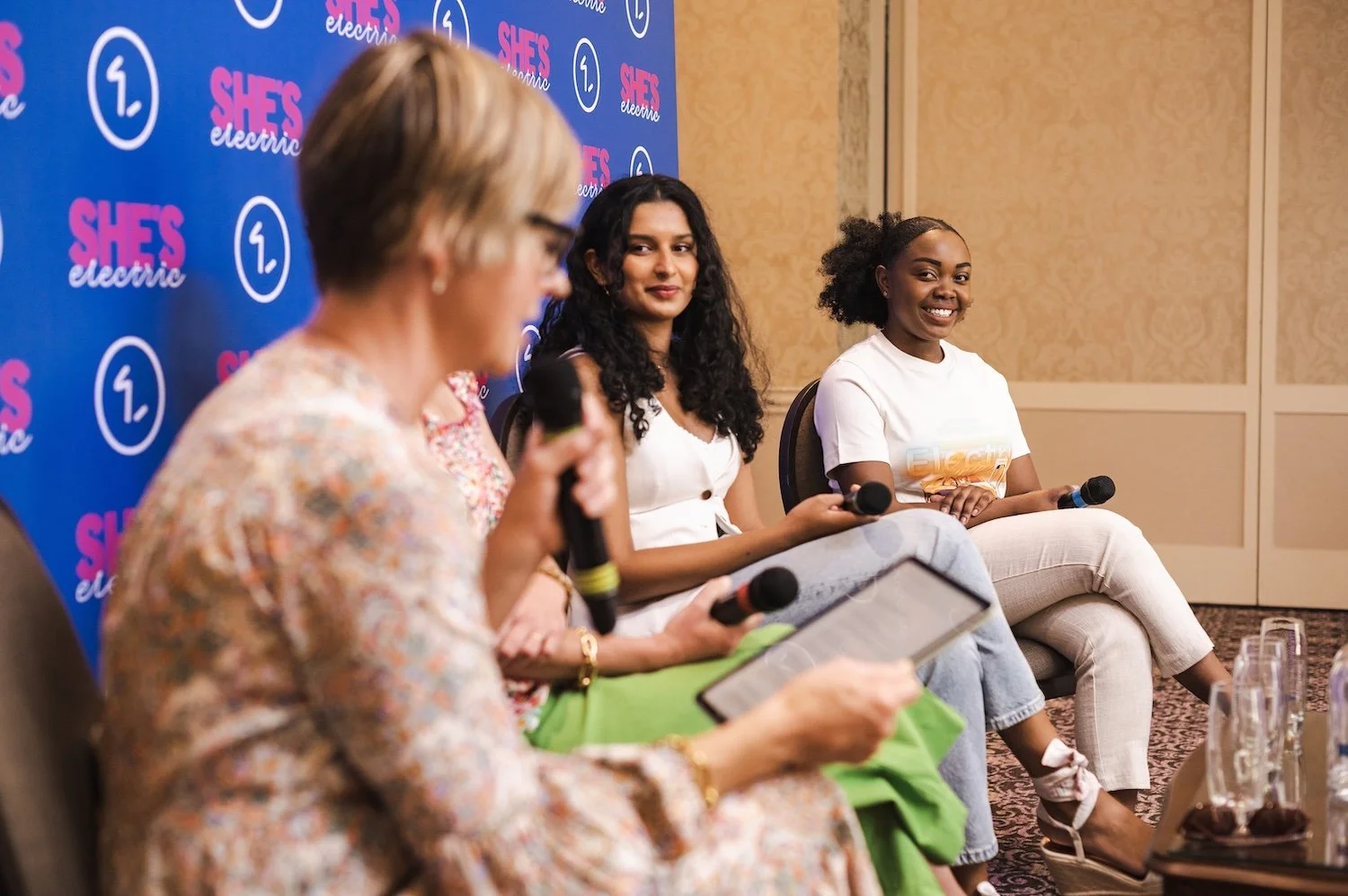 Three women sitting on a panel at an event, with a blue backdrop that has pink and white text reading 'SHE'S electric' and logos.
