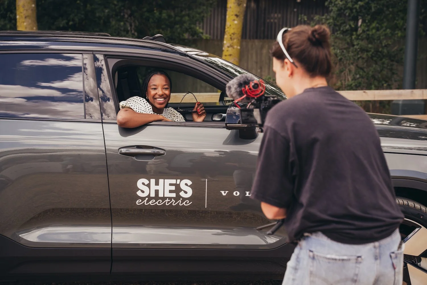 A woman sitting in a car smiling and waving to a filmmaker operating a camera, with a sign on the car that reads "She’s electric".