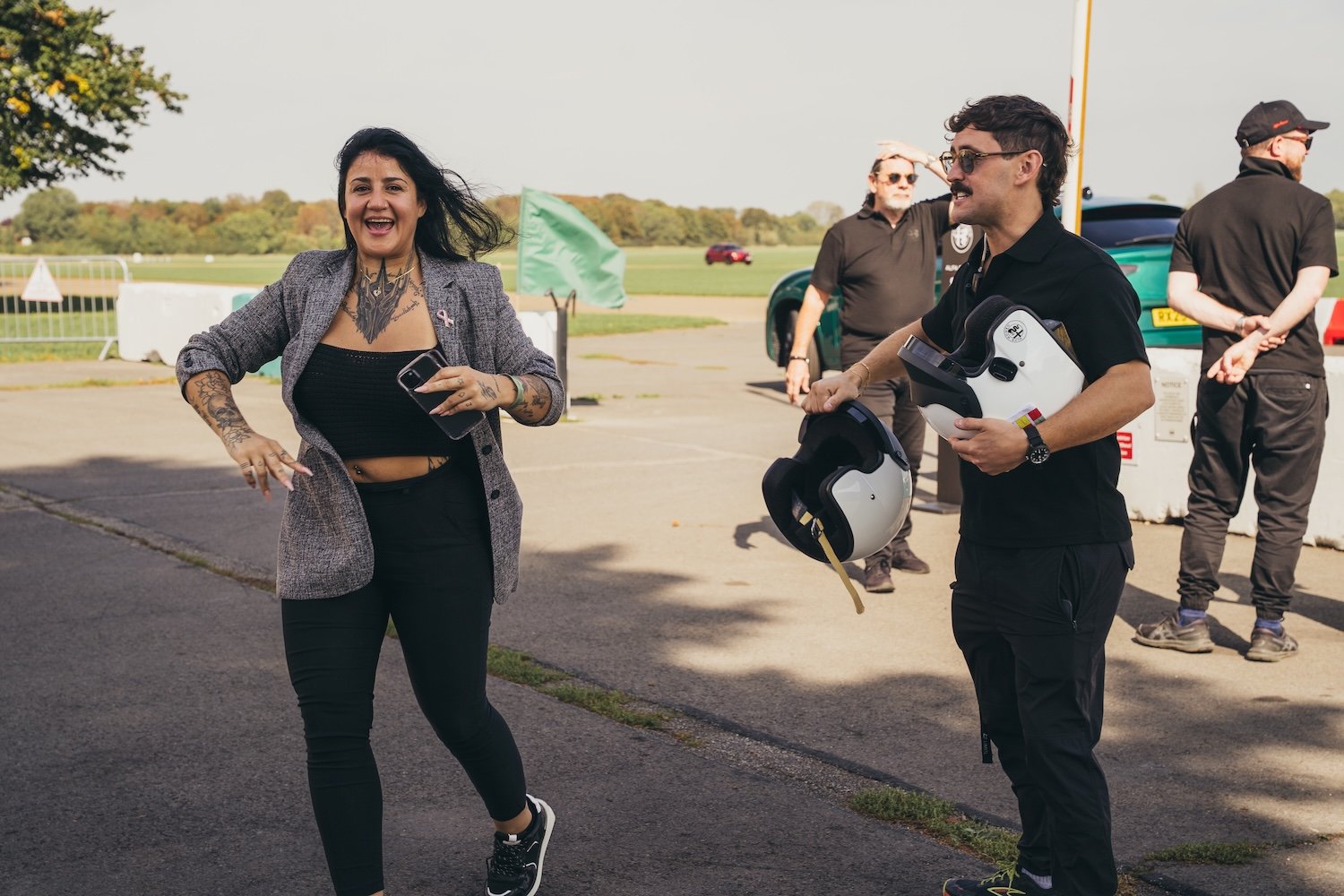 A woman with tattoos on her arms and chest laughing and talking to a man holding a race helmet and a helmet with a head-up display, while others stand in the background outdoors near a racetrack.