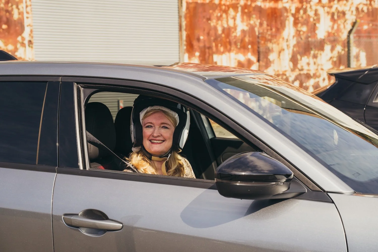 Woman with blonde hair smiling, sitting in the driver's seat of an alfa romeo electric car, wearing a white helmet, with a background of a rust-colored wall 