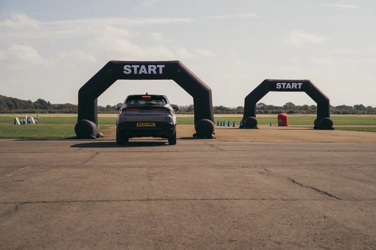 A black Alfa Romeo electric car positioned at a starting line under an inflatable 'START' arch on a wide open flat paved area, with another arch and cones in the background 