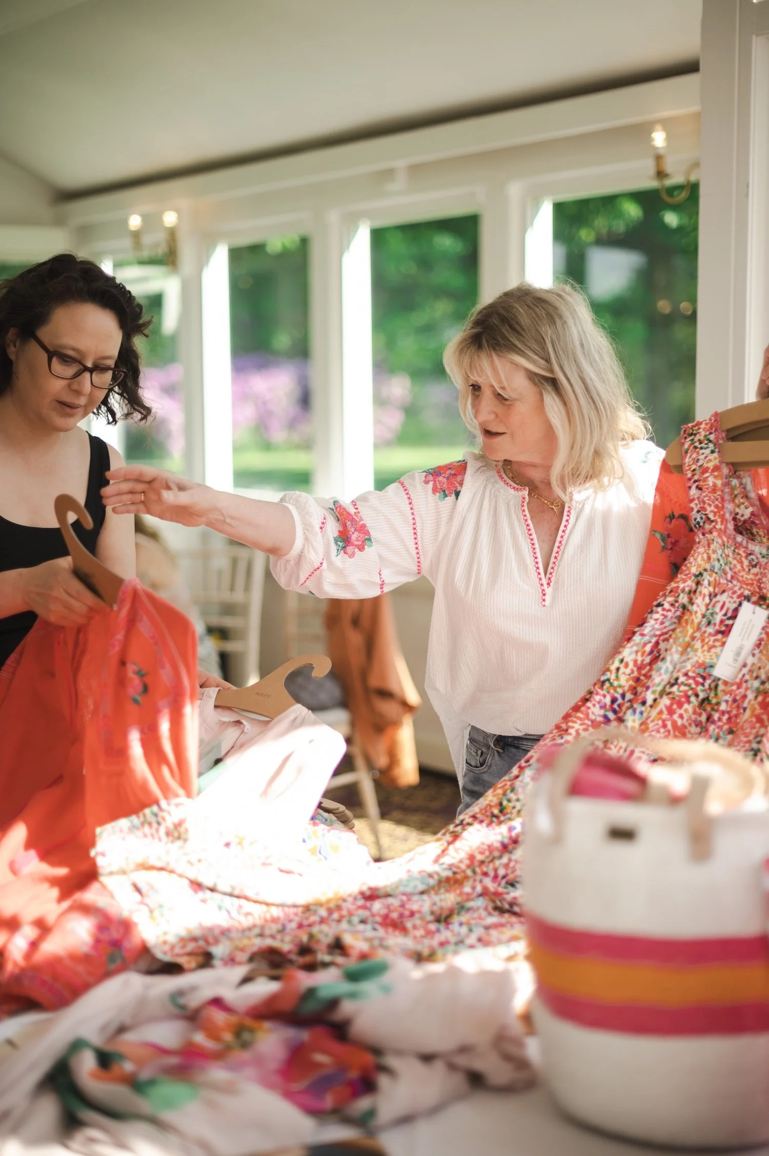 Two women shopping for colorful dresses inside a well-lit room with large windows and a view of green trees outside.