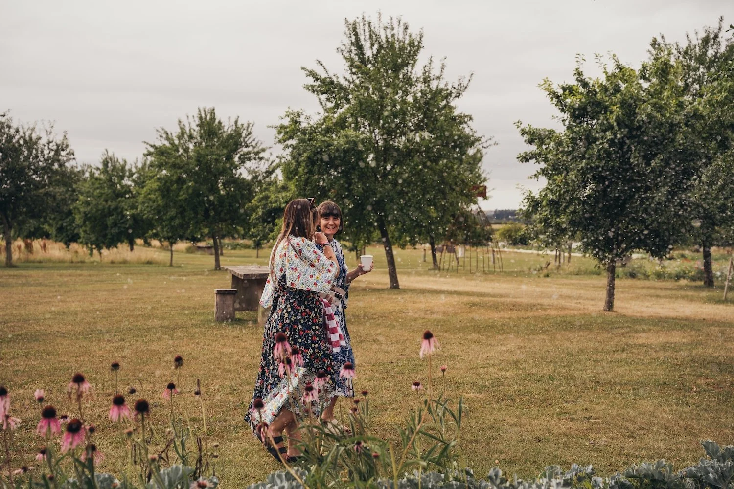 Two women in floral dresses enjoying a walk and conversation in a park with trees and flowers on an overcast day.