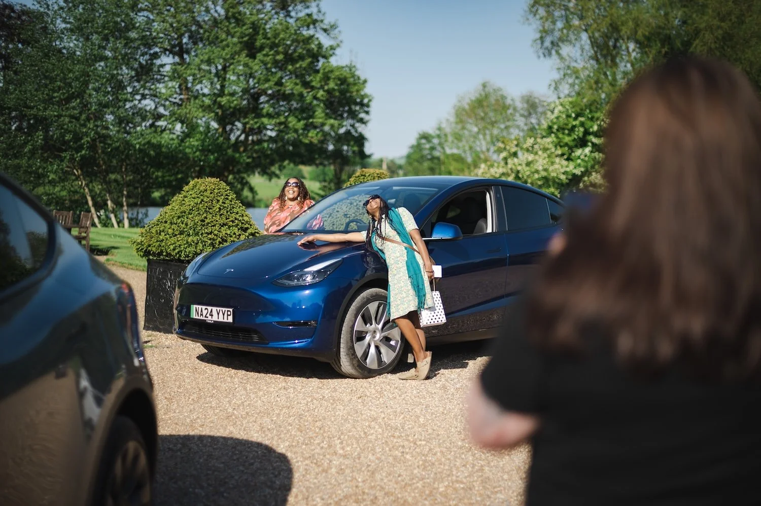 A woman with long braids wearing sunglasses and a dress leaning on the hood of a blue Tesla car, smiling. Another woman with curly hair in a black top, holding a camera, capturing a photo of the scene. The background features a park-like setting with