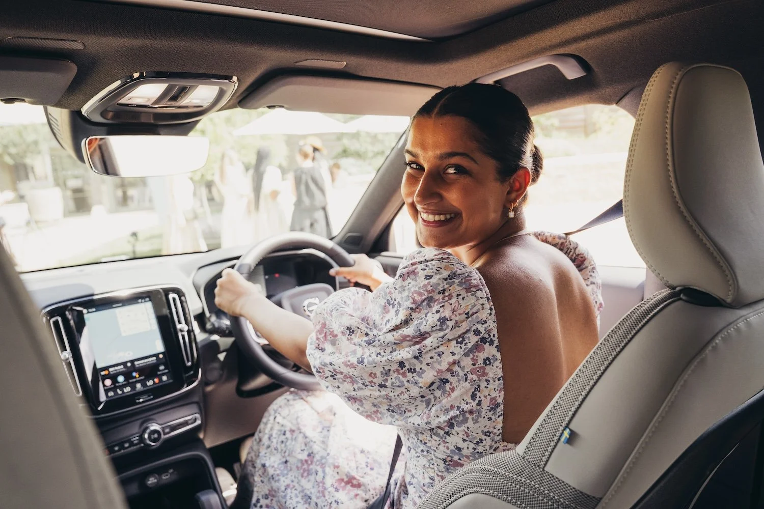 A woman with dark hair smiling inside a car, sitting in the driver's seat and looking back at the camera, with a city street visible through the windshield.