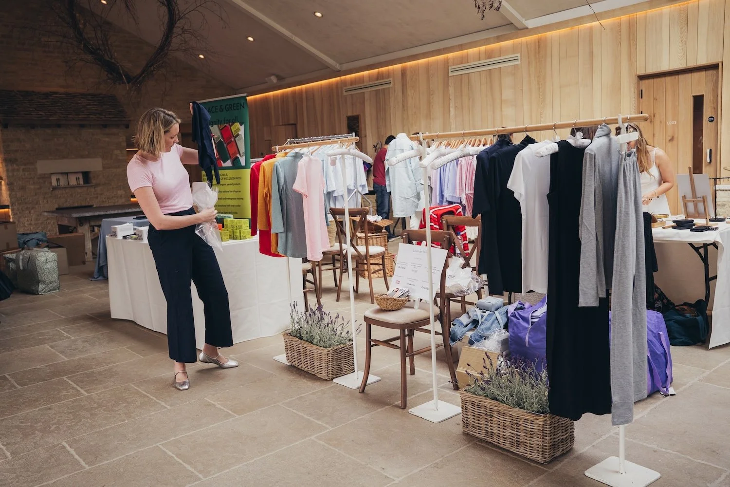 Shopping  at a She's Electric event with racks of colorful clothing, a woman looking at a garment, and another woman behind the table, with plants and chairs in the background.
