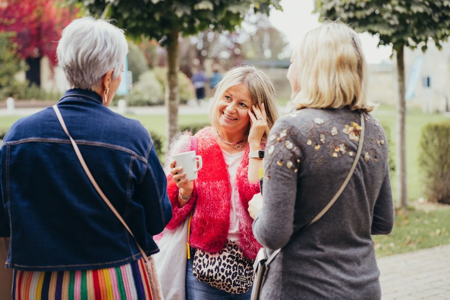 Three women chatting outdoors, one holding a white mug, smiling, with trees and a park in the background.