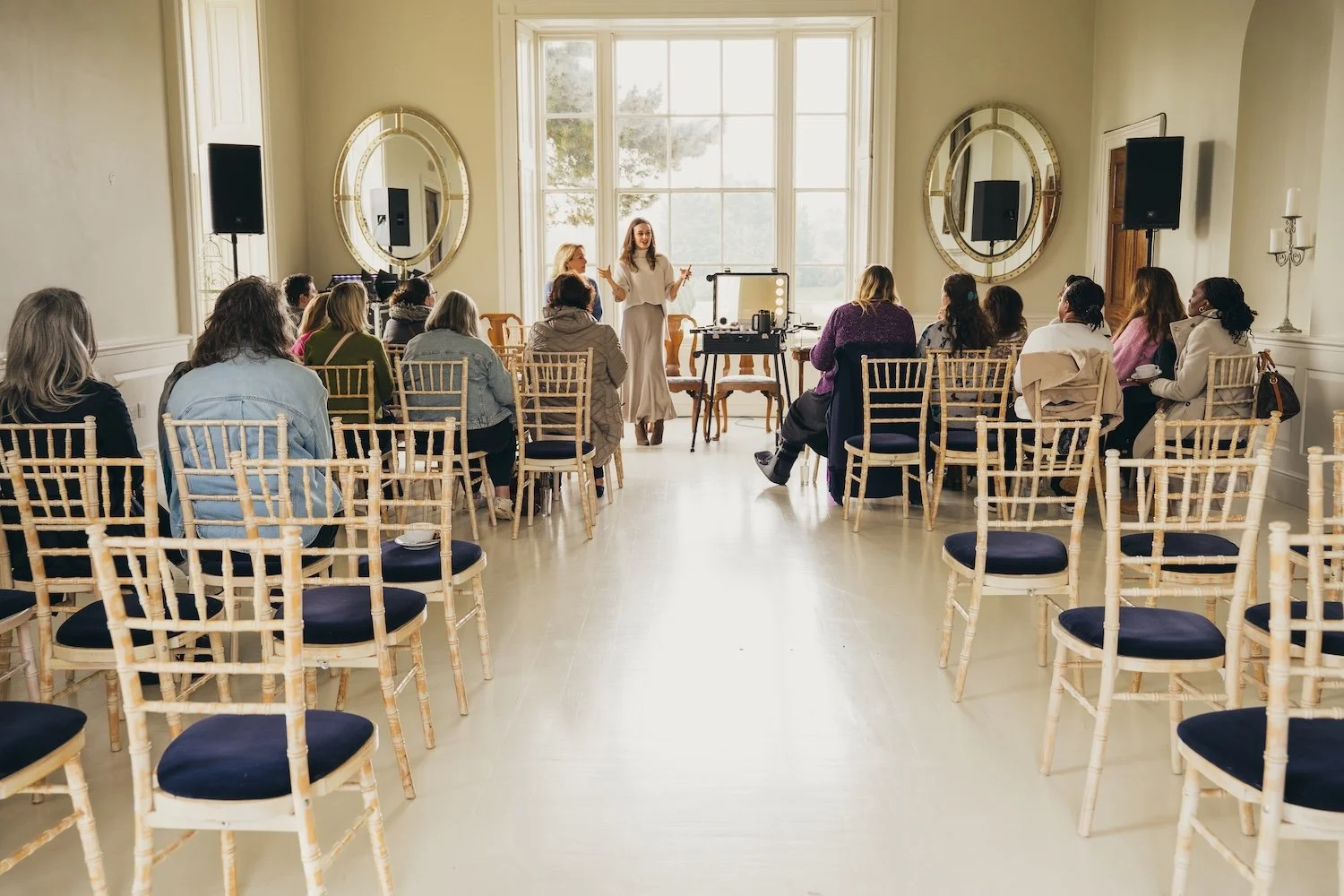 A woman giving a presentation to a seated audience in a bright room with large windows and gold-framed mirrors.