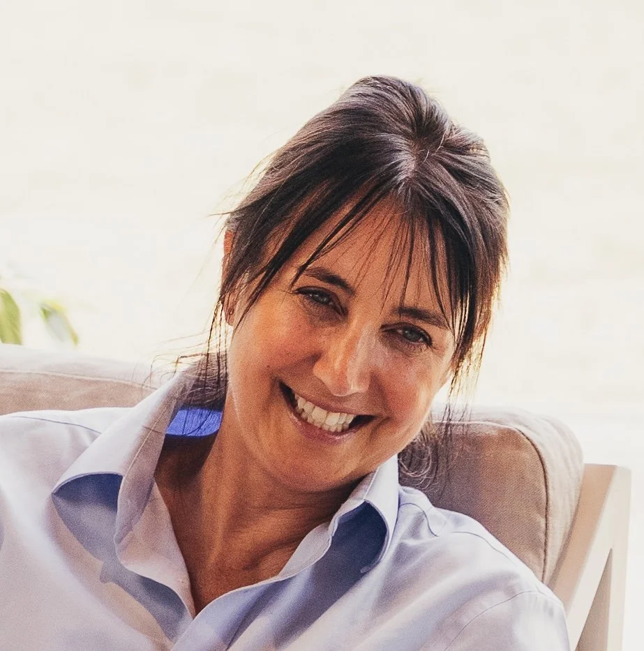 A woman with dark brown hair and bangs smiling and sitting on a beige couch, wearing a light blue button-up shirt.