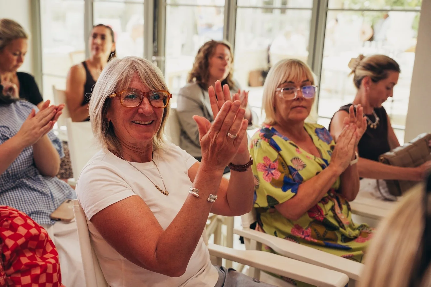 Group of women seated indoors, clapping and smiling, with sunlight coming through glass doors in the background.