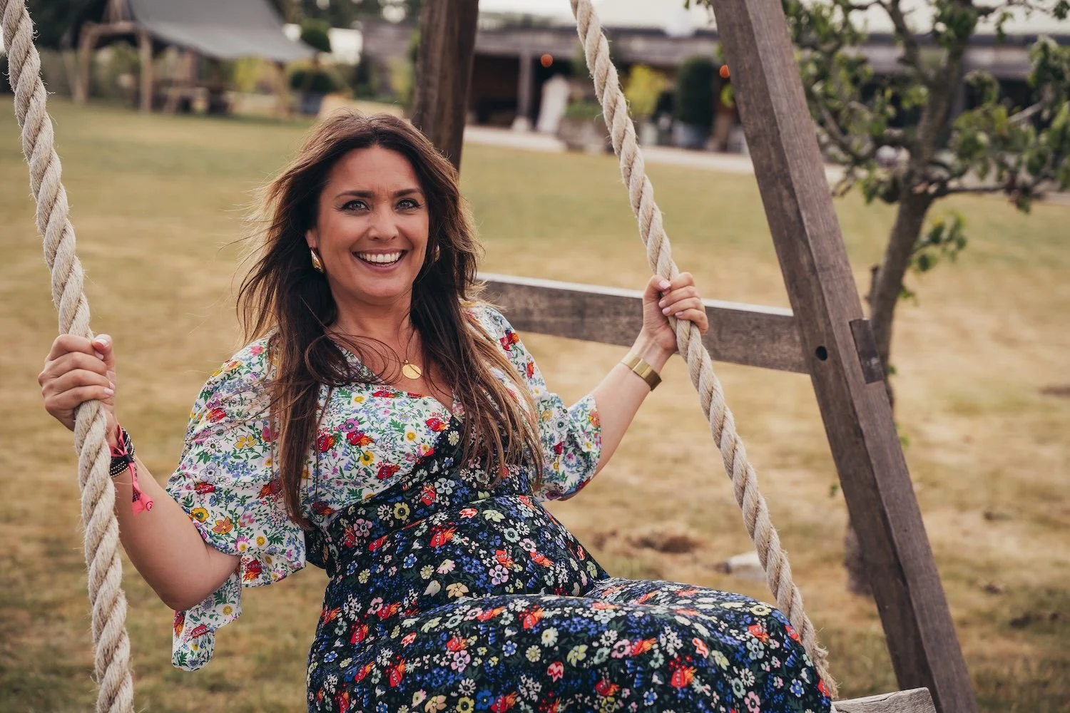 A smiling woman with long brown hair sitting on a wooden swing, wearing a colorful floral dress and gold jewelry, outdoors in a grassy area with trees.