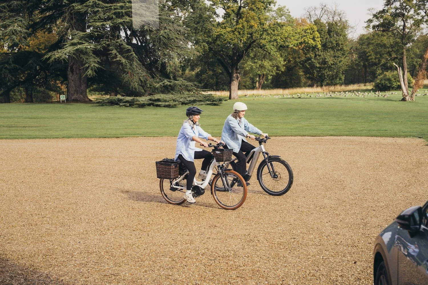 Two women riding e-bikes on a gravel path in a park with trees and grass in the background at a She's Electric event