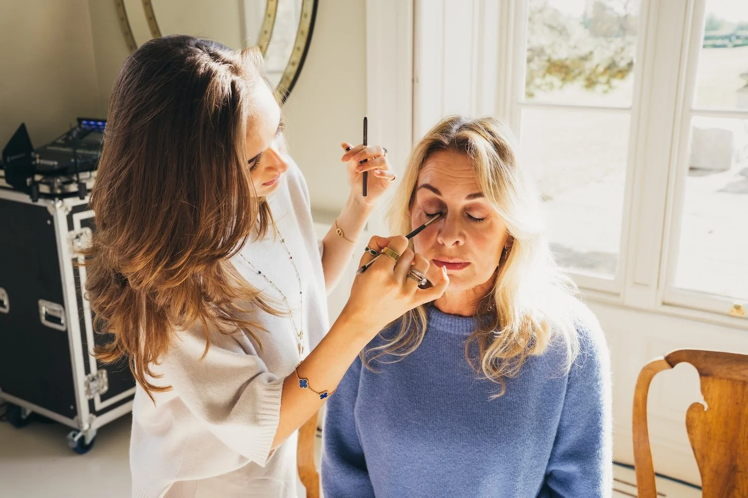 A woman applying makeup to another woman sitting on a chair near a window.