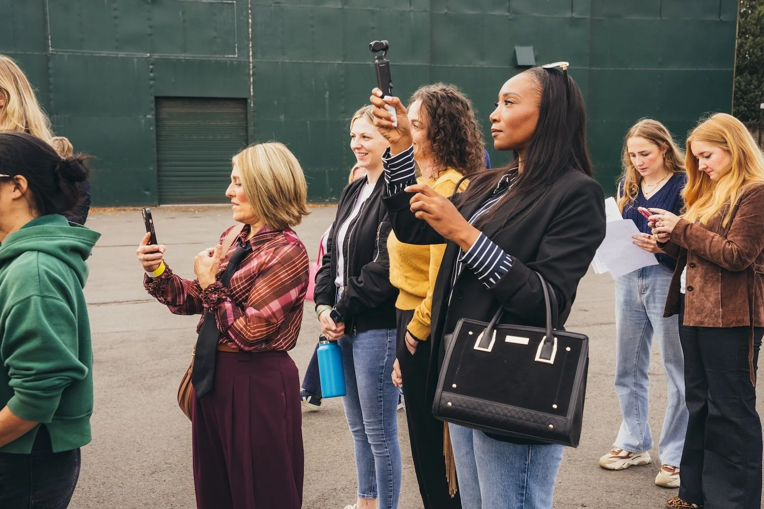 A group of women standing outdoors, some taking photos or looking at their phones