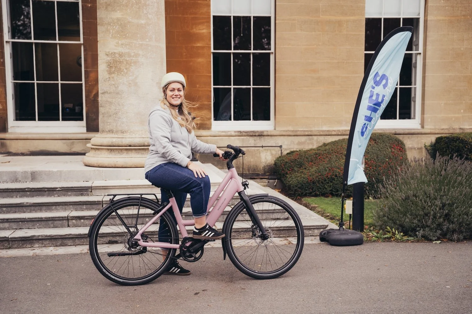 A woman wearing a helmet sitting on a pink e-bike in front of a building with steps and large windows, with a blue promotional she's electric flag nearby.